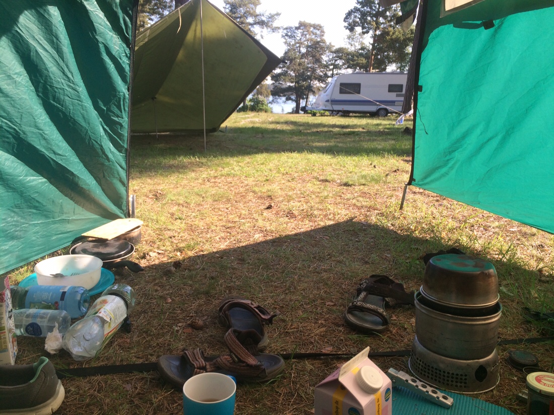 Blick aus dem Zelt mit Sandalen, Teetasse, vorne das Tarp, am Horizont Bäume und Meer