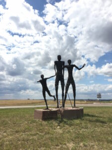 Drei menschliche Skulpturen aus Draht auf roten Betonsockeln, auf Wiese unter wolkigem Blauhimmel: ein Kind und zwei Erwachsene