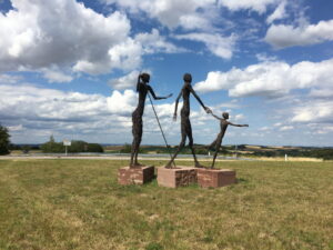 Drei menschliche Skulpturen aus Draht auf roten Betonsockeln, auf Wiese unter wolkigem Blauhimmel: ein Kind und zwei Erwachsene