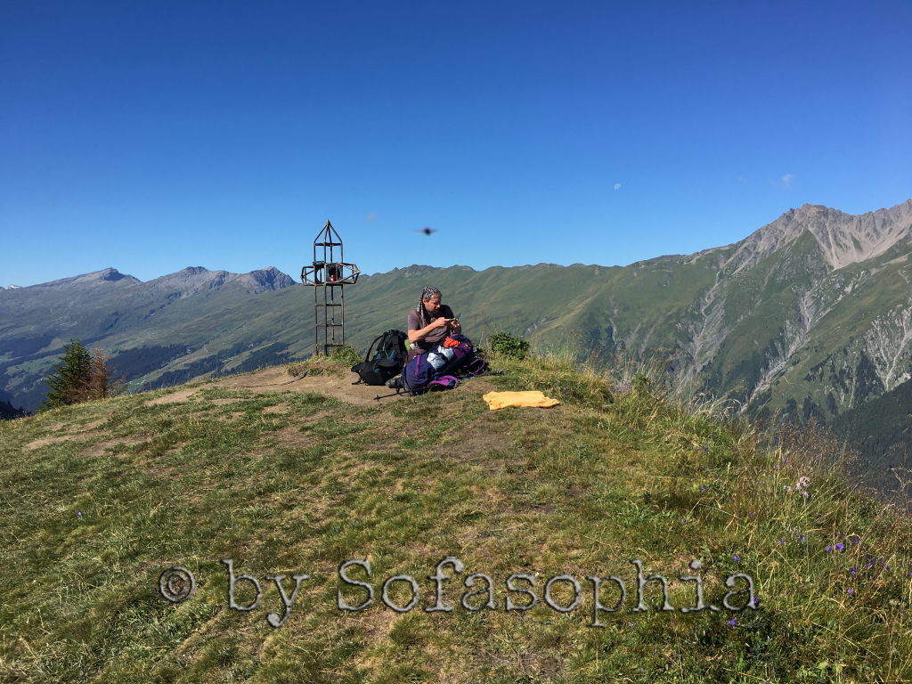 Irgendlink sitzt auf dem Tguma am Boden auf der Wiese und betrachtet seine Aufnahmen im Handy. Hinter ihm das Gipfelkreuz.