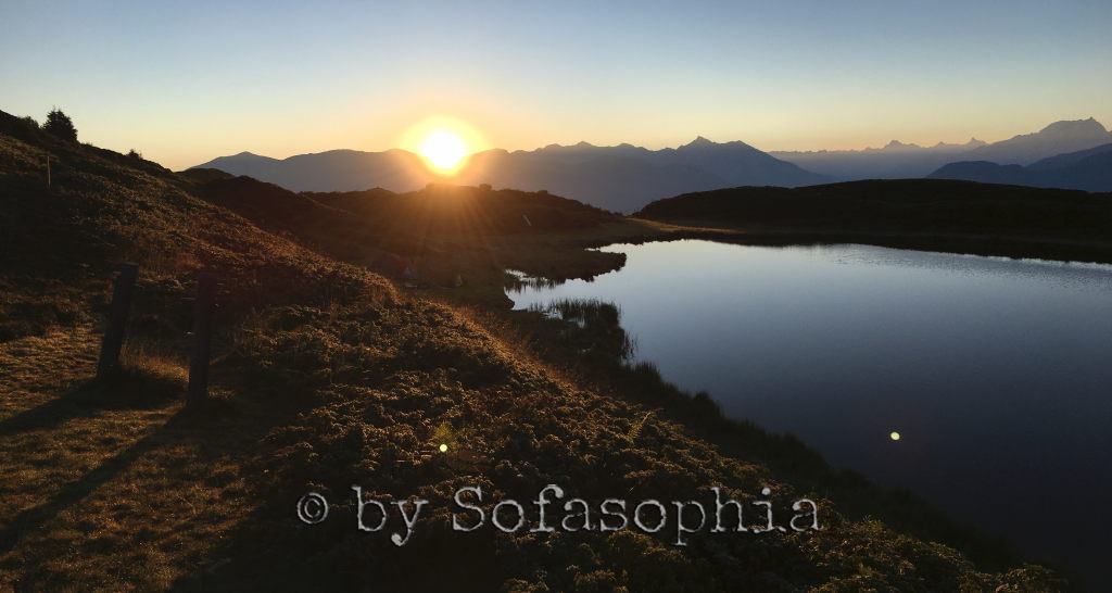 Sonnenaufgang vom Hügel oberhalb des Sees. Die Sonne taucht alles in goldenes Licht, der See glänzt.