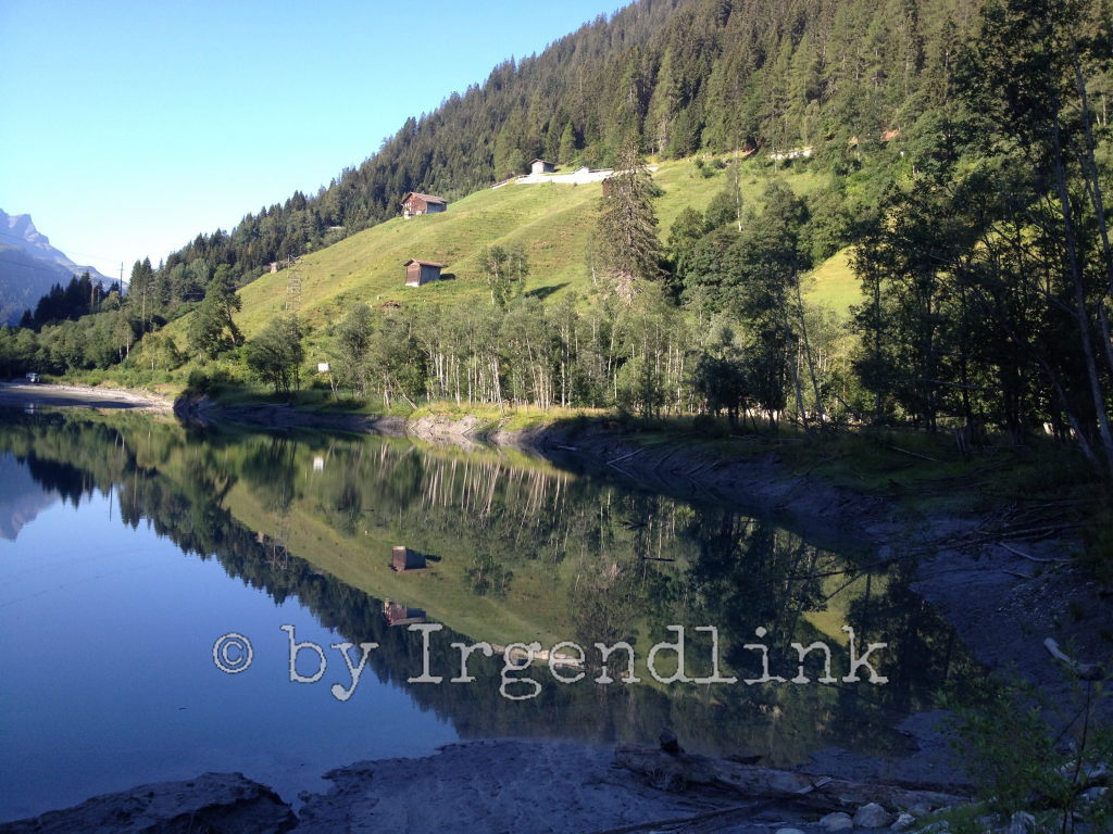 Am Stausee ein Blick in die Hügel. Alphütten auf den Wiesen. Darüber Wald. Alles spiegelt sich im See.