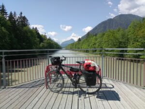 Reiserad geleht an rote Metallbank auf Brücke, vor Fluss und Bergen im Hintergrund, darüber Blauhimmel