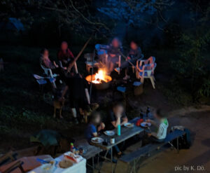 hinten links eine Gruppe Menschen an einem Lagerfeuer, vorne Menschen an einem Tisch, die UNO spielen. Die Gesichter sind unscharf