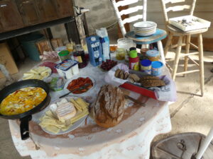 Frühstücksbüffet auf rundem mit Tischtuch bedecktem Tisch und auf Stühlen, Brot, Käse, Hafermilch etc.