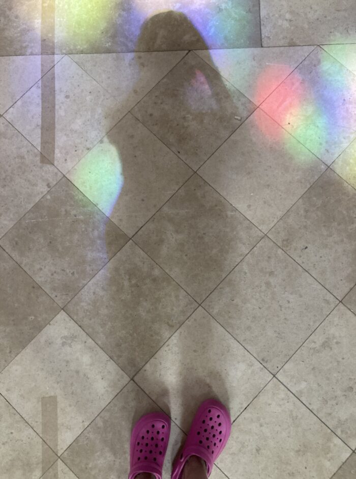 Hellgrauer Plattenboden mit dem Schattenriss einer Person. Der Boden reflektiert bunte Flecken, die von bunten Fenstern stammen. Am unteren Bildrand sind pinke Crocs zu sehen.