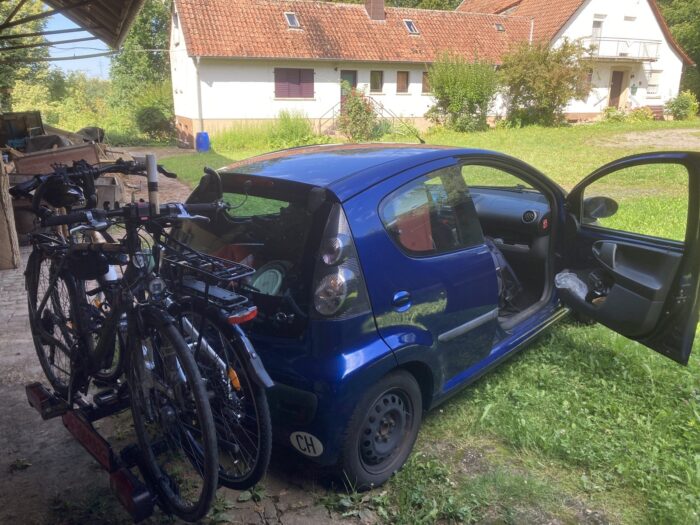 Vollgepacktes kleines blaues Auto vor der Abreise. Im Hintergrund eine Wiese und ein längliches Gebäude.