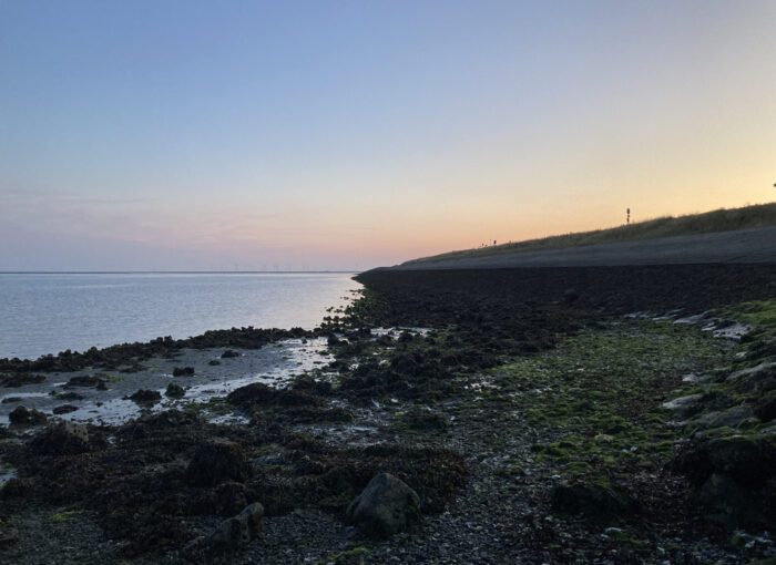 Ruhige Strandaufnahme bei Sonnenuntergang: links Meer, rechts Ufer, steiniger Strand, ansteigendes Gelände.