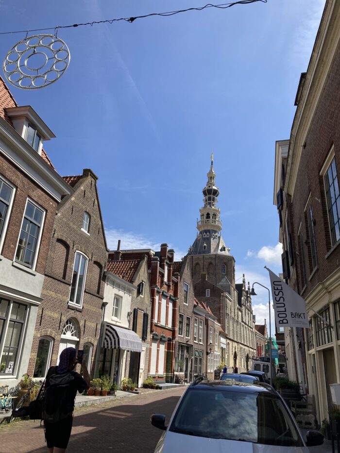 Stadtaufnahme. Alte Häuserfassaden, in der Bildmitte ein zwiebelartiger Kirchturm. Im Vordergrund der Schatten eines fotografierenden Menschen.