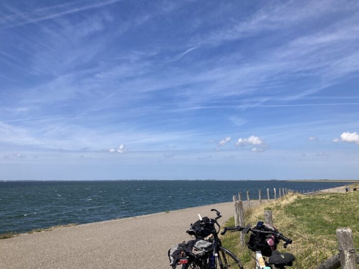 Im Vordergund zwei Fahrräder, im Hintergrund Meer und Blauhimmel, rechts der Radweg mit Zaun und Wiese.