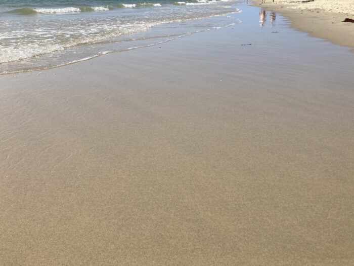Nasser Sand und eine anrollende Welle. Im Hintergrund spiegeln sich zwei Menschen im Wasser. Rechts ein wenig trockener Sand.