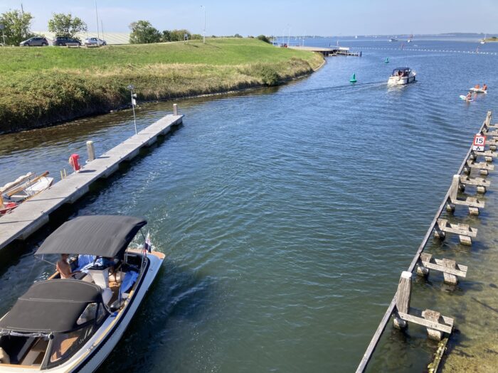Hafenszene. Zwei Motorboote fahren auf einem Kanal, der auf das Meer hinausfließt. Links grünes Uferland, rechts eine metallene Vorrichtung als Abgrenzung zwischen Wasser und Land.