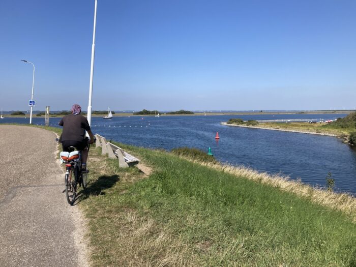 Radelnder Mensch von Hinten auf einem Radweg, der direkt am Kanal entlang führt. Der Kanal mündet weiter vorn, in der Bildmitte, ins Meer.