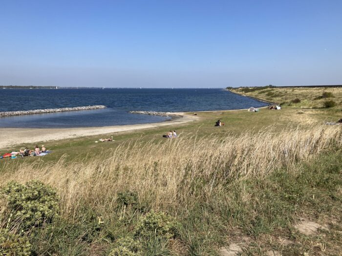 Badestrand von oben. Im Vordergrund hohes Gras. Darunter kurzgeschnittene Liegewiese, Sandstrand und Meer. Einige Menschen lagern auf der Wiese. Blauhimmel.