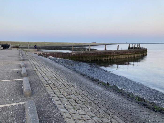 Meer, betonierter Strand mit Kies, Mauern, Landzunge – bei untergehender Sonne.