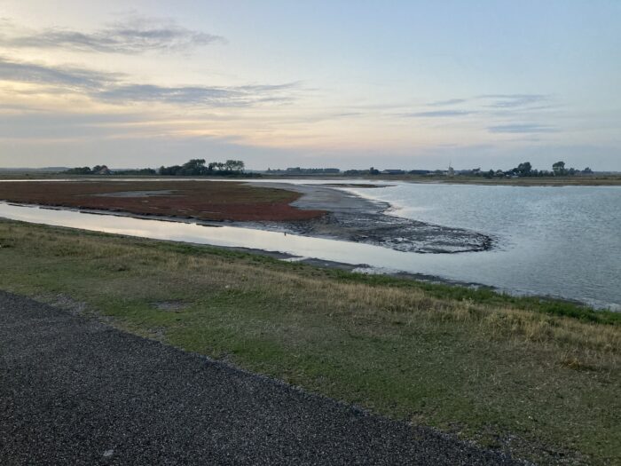 Blaue Stunde. Orange-farbener Himmel mit dunkelgrauen, schlierigen Wolken. Gewässer auf der rechten Bildseite, darum herum festes Land, im Vordergrund ein grasbewachsener Abhang.