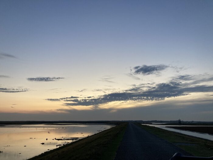 Blaue Stunde. Orange-farbener Himmel mit dunkelgrauen, schlierigen Wolken. Gewässer auf der rechten und linken Bildseite, in der Mitte, im Vordergrund eine Art Deich mit Radweg.