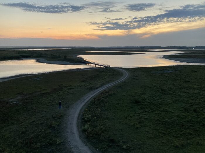 Sonnenuntergang. Ausblick von Aussichtsturm über Gewässer, eine Brücke in der Bildmitte, bewölkten Himmel und im Vordergrund ein Radweg, der sich grau vom restlichen Land abzeichnet.