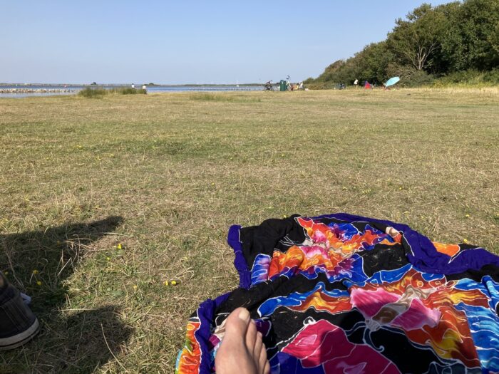 Strandwiese, im Hintergund Bäume, Meer und ein paar wenige Menschen am Strand. Im Vordergrund ein Fuß, ein buntes Baumwolltuch und Schatten.