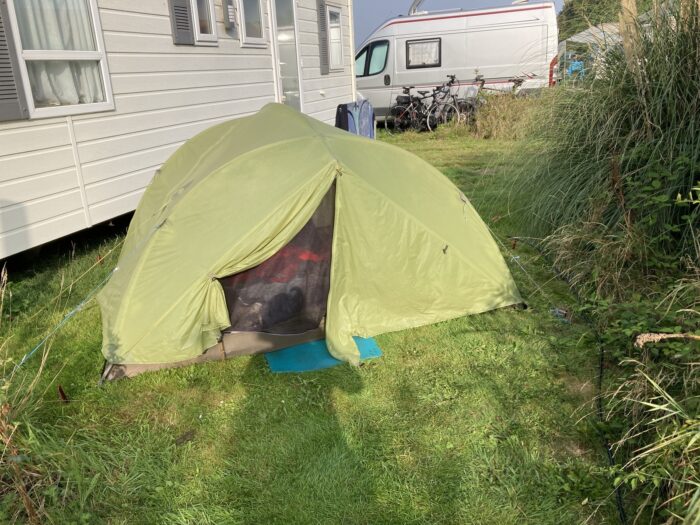 Hellgrünes Zelt neben weißem Mobilhome, im Hintergrund Wohnmobil. Im Vordergrund Rasen und der Schatten einer Person.