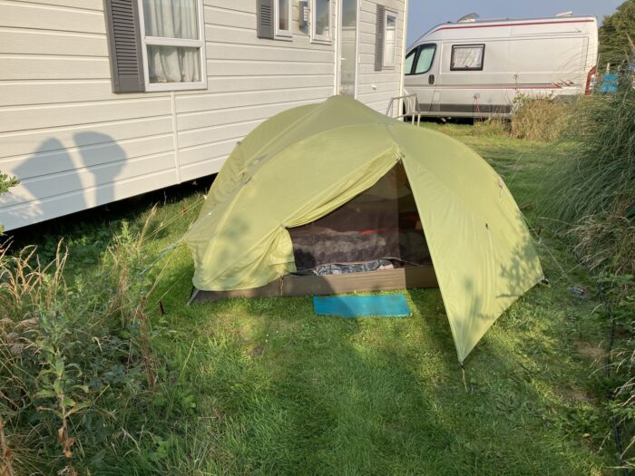 Tisch-Bänke-Garnitur in der Bildmitte. Auf dem Tisch stehen Campingsachen, davor ein kleines Tischchen mit einem Kocher drauf. Im Vordergrund eine Wäscheleine mit Tüchern. Der Tisch steht auf einem Holzboden. Im Hintergrund sind Hecken und Bäume, rechts ein Stück des Mobilhomes.