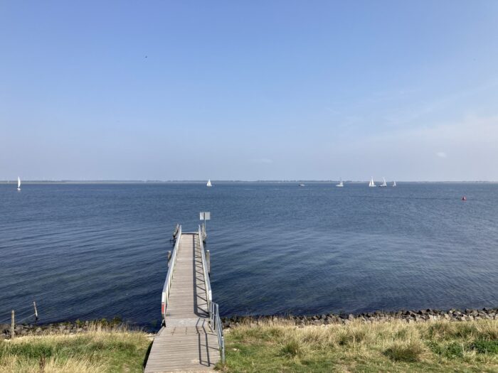 Steg ins Meer, im Hintergrund Meer und am Horizont ein paar Boote. Darüber blauer Himmel. Im Vordergrund eine Wiese.
