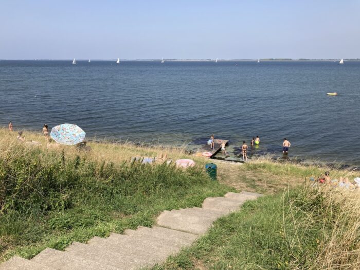 Blick vom Deich aus auf eine steinerne Treppe, die an einen schmalen Strand führt. Ein Sonnenschirn, dazu ein paar Leute – im Wasser und an Land. Meer, einige Boote, blauer Himmel.