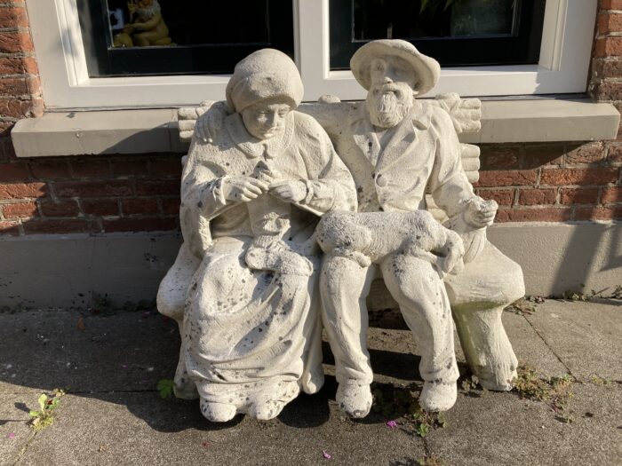 Steinskulptur vor einem Backsteinhaus mit weißem Fenster. Die Skulptur zeigt ein älteres Paar, das auf einer Bank sitzt. Die Frau strickt, der hut- und barttragende Mann sitzt daneben.