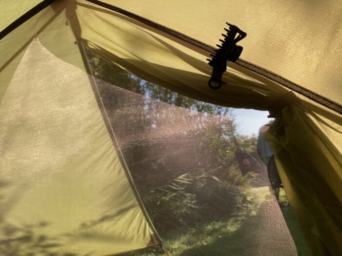 Morgendlicher Blick aus dem Zelt. Die äußere Zelthaut ist aufgeklappt, das innere Netz ist geschloßen, das Sonnenlicht wirft Muster auf das Netz. Am Zelt klemmt innen eine schwarze Haarklammer.