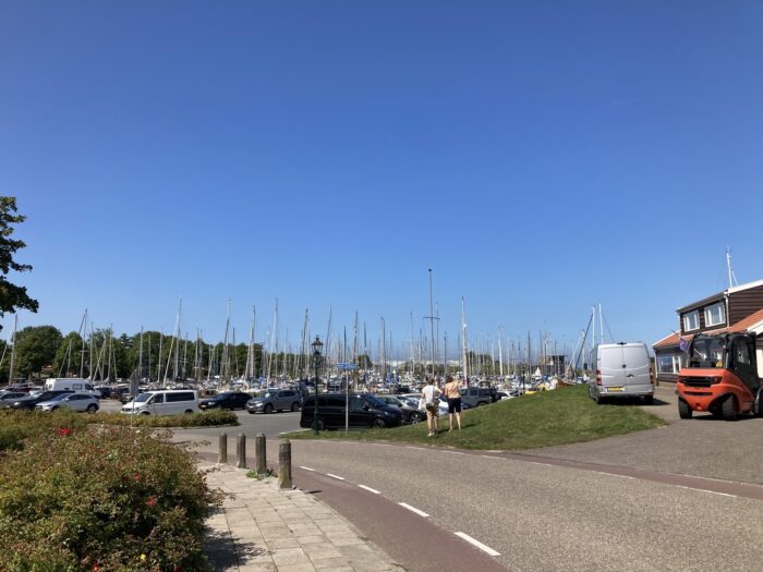 Hafenszene mit vielen Masten im Hintergrund, im Vordergrund eine Straße mit Radweg am linken Rand und Gehweg für Fußgänger*innen. Blauhimmel.