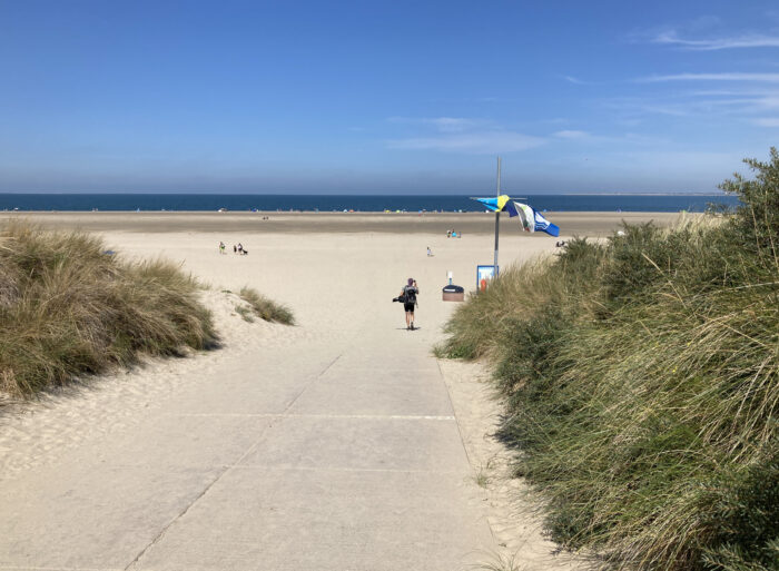 Sandstrand im Weitwinkel. Von oben, vom Deich aus, fotografiert. Das Meer ist quer in der Bildmitte als dunkelblauer Streifen zu sehen. Vorne Sand und links und rechts Gebüsch. Darüber Blauhimmel.