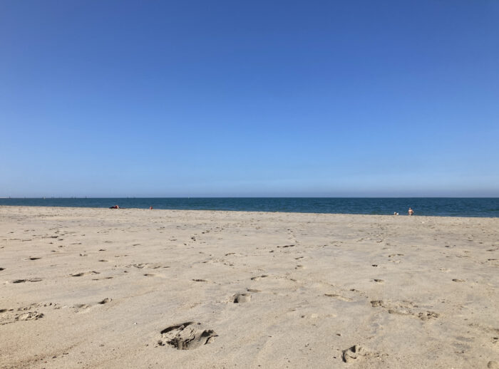 Vorne Sand, in der Mitte ein Streifen Meer und weit entfernt zwei Menschen, darüber sattblauer Himmel.