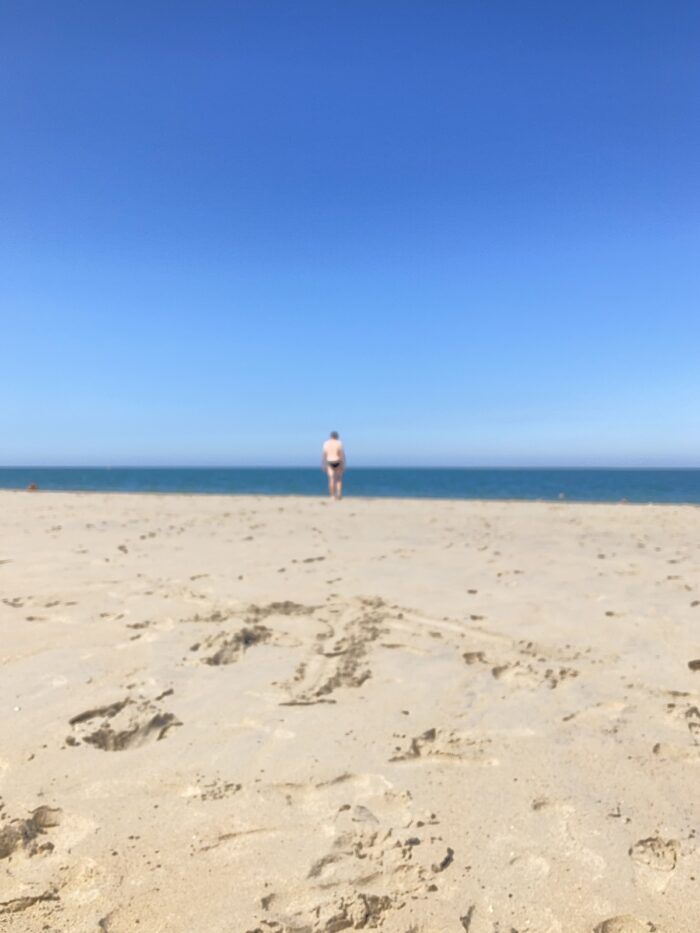 Vordergrund Sand mit Fußspuren, in der Mitte ein Streifen Meer und ein unscharf fotografierter Mensch in Badehose, darüber sattblauer Himmel.