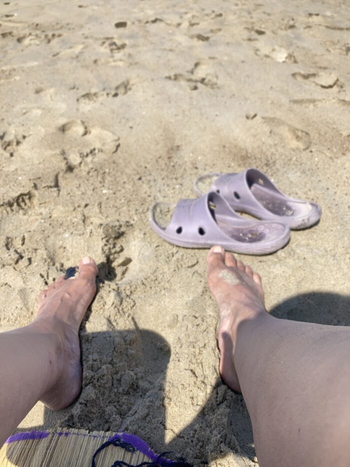 Füße und Unterschenkel im Sand, lila Schlappen, Sand, ein Stück Bastmatte vorne links.