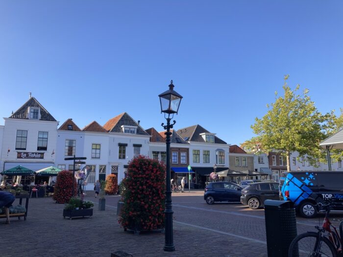 Dorfplatz mit alten, einstöckigen Häusern, einer Straßenlaterne, Autos, Fahrrädern und Bäumen. Blauer Himmel, der nach Spätnachmittag aussieht.