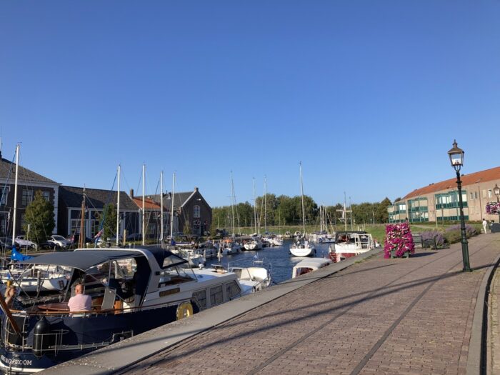 Hafenszene auf dem Kanal. Schiffe, viele Masten, rechts das Ufer mit einer Straßenlaterne, links das andere Ufer mit Häusern. Im Hintergrund Bäume, darüber ein blauer Himmel.