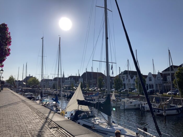 Sonne am blauen Himmel, darunter der Kanal mit Schiffen, im Hintergrund, am anderen Ufer Häuserzeile, im Vordergrund Straße am Ufer.