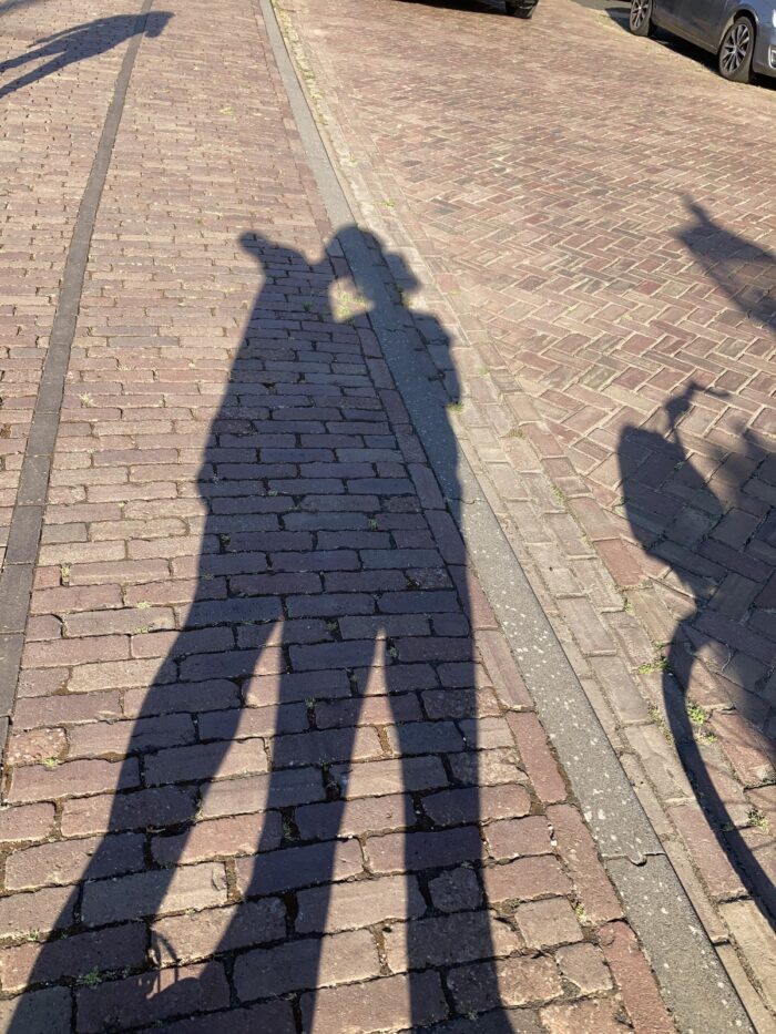 Schattenselfie zweier Menschen auf Pflastersteinstraße, drumherum weitere Schatten ... rechts der Schatten eines Fahrrades.