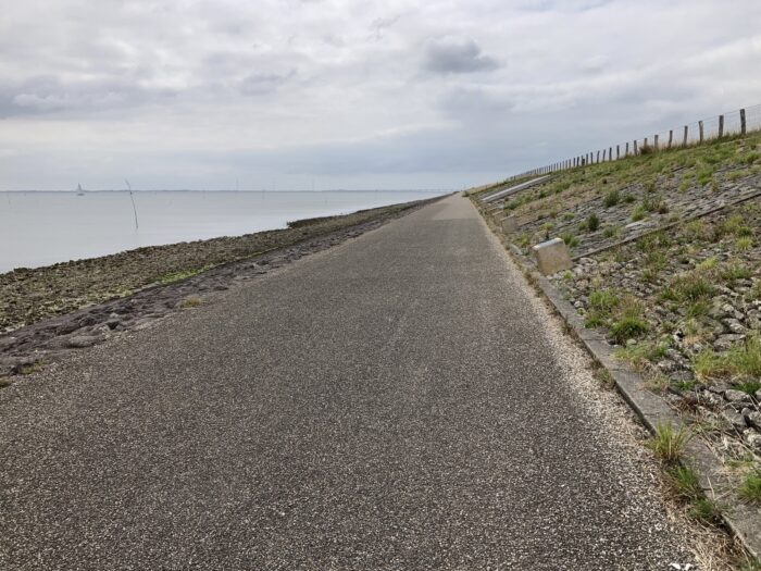 Radweg von vorn nach hinten verlaufend. links das Meer an einem abschüssigen steinigen Hang, rechts Deichmauer. Der Himmel ist bewölkt und grau.