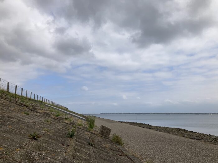 Dramatischer Himmel mit weißen und grauen Wolken über Radweg und Deich, auf der rechten Seite das Meer.