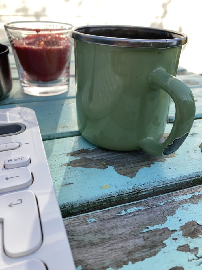 In der Bildmitte eine türkisfarbene Emailletasse auf einem Holztisch, im Hintergrund eine rote Kerze in einem durchsichtigen Glas, vorne ein Stück einer weißen Tastatur.