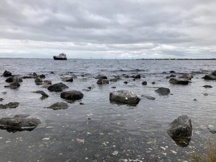 Meeraufnahme, kiesiger steiniger Strand mit großen Steinen, am Horizont ein Motorschiff, darüber ein dramatischer wolkiger Himmel.