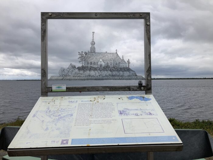 Das Bild zeigt eine Informationstafel an einem Seeufer. Auf der Tafel ist eine Zeichnung einer Kirche auf einer Insel zu sehen, die vermutlich eine historische Darstellung eines versunkenen Dorfes ist. Der Text auf der Tafel beschreibt die Geschichte dieses Dorfes, „Bommee“, und seinen Untergang. Der Hintergrund des Bildes ist ein trüber Himmel und ruhiges Wasser. Die Tafel dient der Information und Erinnerung an die verlorene Siedlung. Da der Bildrahmen mit der Kirche durchsichtig ist und der Aufnahmewinkel so gewählt ist, dass der untere Rand der Kirche auf den Horizont des Meeres im Hintergrund trifft, sieht es aus, als ob die Kirche auf dem Meer steht.