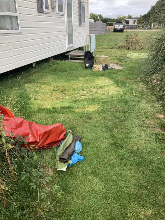 Wiese nun ohne Zelt, da wo das Zelt stand, ist das Gras gelb verfärbt. LInks das Mobilheim, vorne Taschen und grüne Wiese.