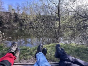 Drei bewanderschuhte Beinpaare nebeneinander auf Holzliege, im Hintergrund der See mit Gewächs