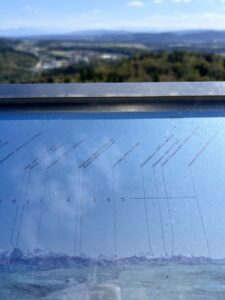 ein sogenannter Alpenzeiger, eine Metalltafel, die das Bergprofil mit Beschriftungen der Berge in der Ferne abbildet, dahinter die Alpen den Berner Oberlandes unscharf