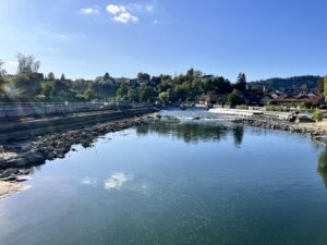 Eine Flusslandschaft mit Blauhimmel- und Wolken-Spiegelungen im Wasser, im HIntergrund Ufer mit Bäumen und Häusern, links das Wehr