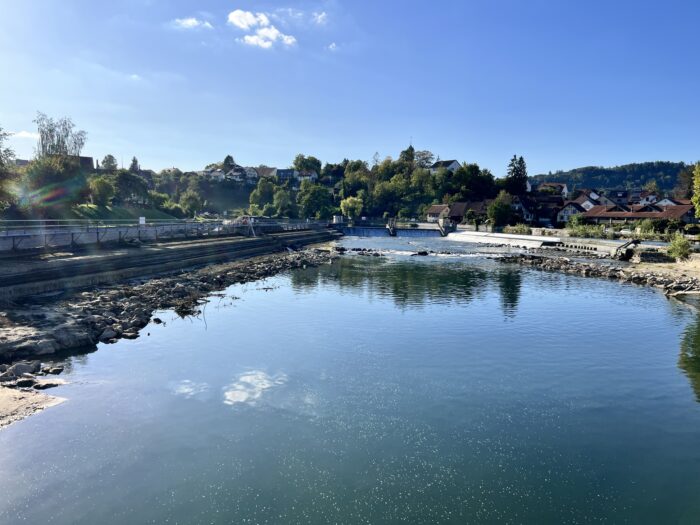 Eine Flusslandschaft mit Blauhimmel- und Wolken-Spiegelungen im Wasser, im HIntergrund Ufer mit Bäumen und Häusern, links das Wehr
