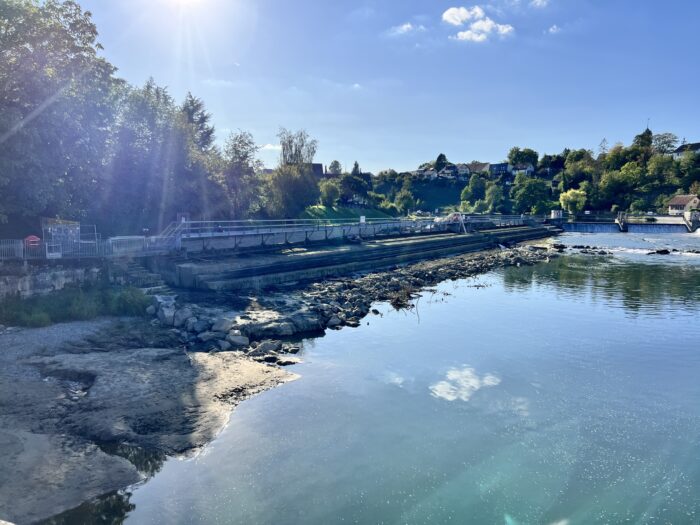 Eine Flusslandschaft mit Blauhimmel- und Wolken-Spiegelungen im Wasser, im HIntergrund Ufer mit Bäumen und Häusern, auch ein Wehr ist zu sehen