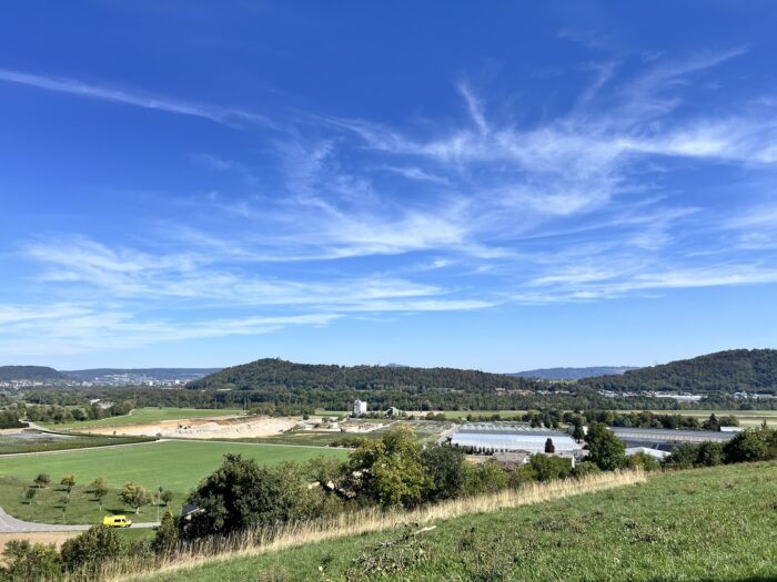 Weitsicht über hügeliges Land, darunter in einiger Entfernung Treibhäuser, darüber Blauhimmel